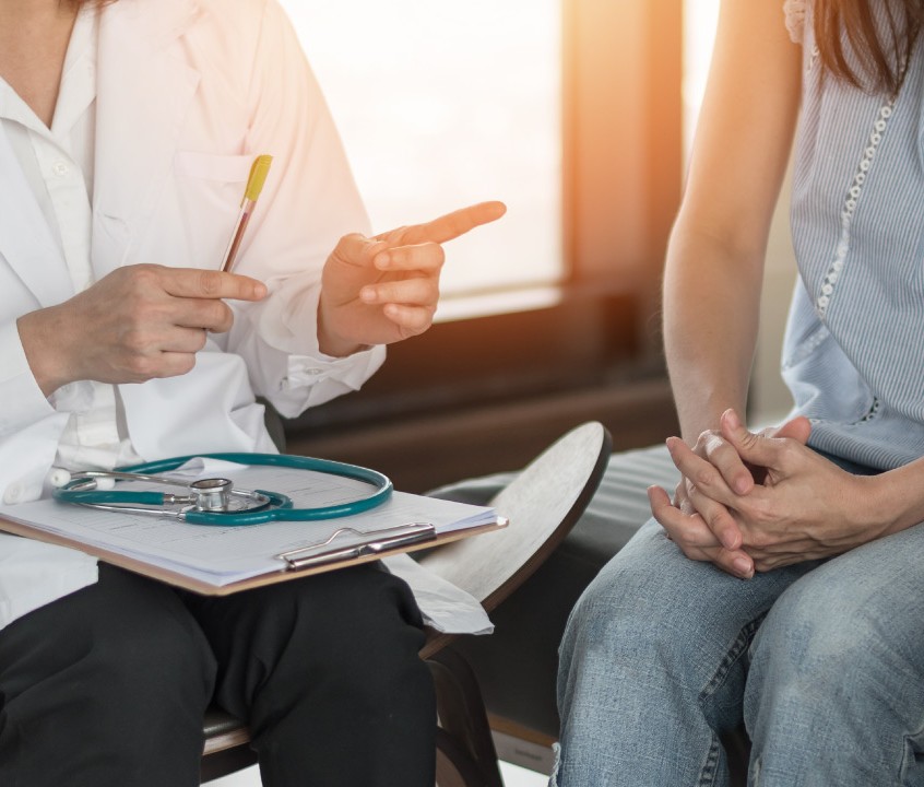 A doctor gestures while discussing with a patient, who holds her hands together, in a bright, modern healthcare setting.