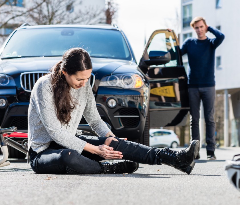 A person sits on the road, appearing injured, while a car door is open nearby and another person stands, looking concerned.