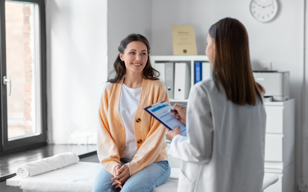 A woman sits on an examination table in a medical office while a healthcare professional discusses information from a clipboard.