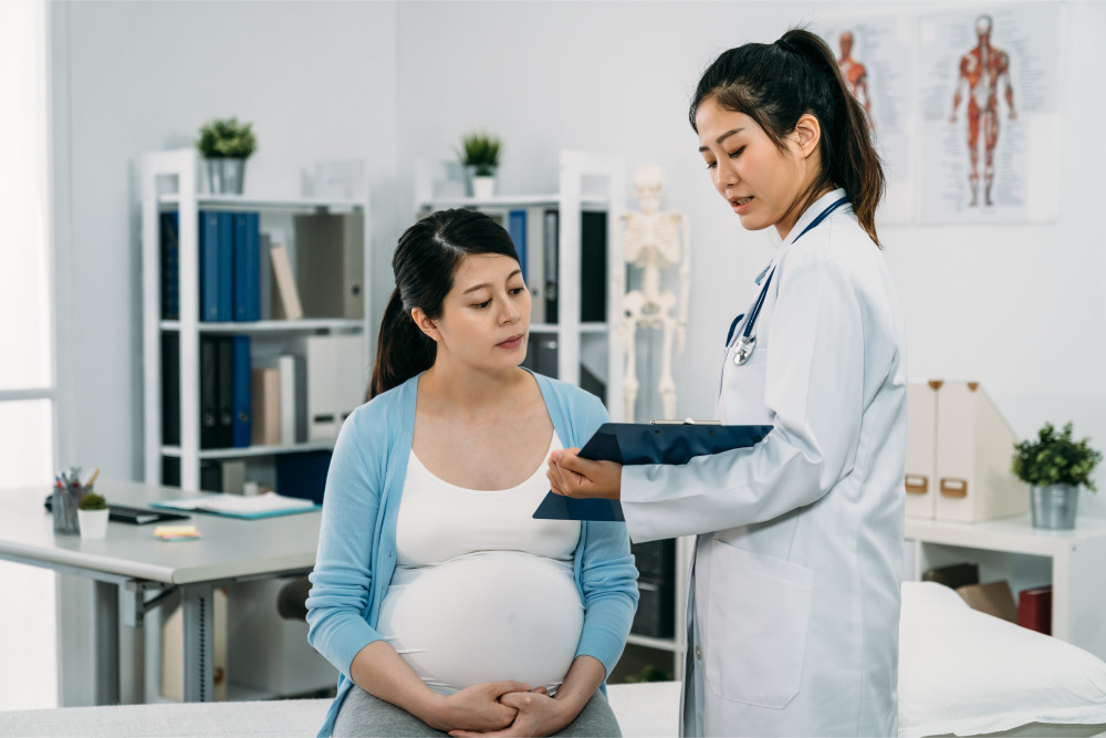 A pregnant woman in a light blue cardigan sits in a doctor's office while a doctor discusses information holding a clipboard.