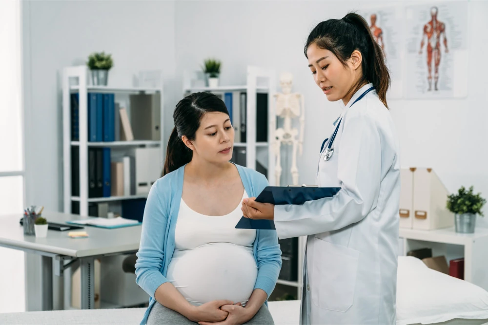 A pregnant woman in a light blue cardigan sits in a doctor's office while a doctor discusses information holding a clipboard.